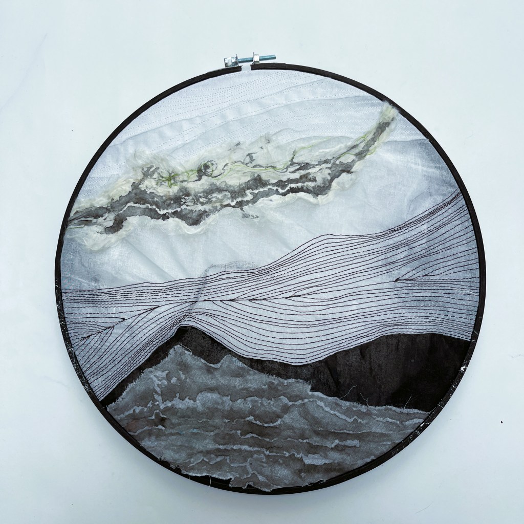 A photo of a multimedia artwork looking like a landscape in a circular painted embroidery frame